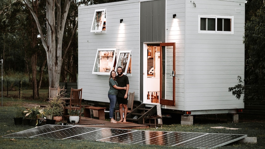 A young couple stand smiling in front of a very small, squarish wood house on cinder blocks. Solar panels sit in the foreground.