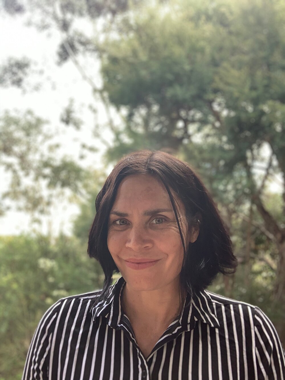 A woman with shoulder length dark wears monochrome vertical striped button up shirt, behind her are lush trees on overcast day.