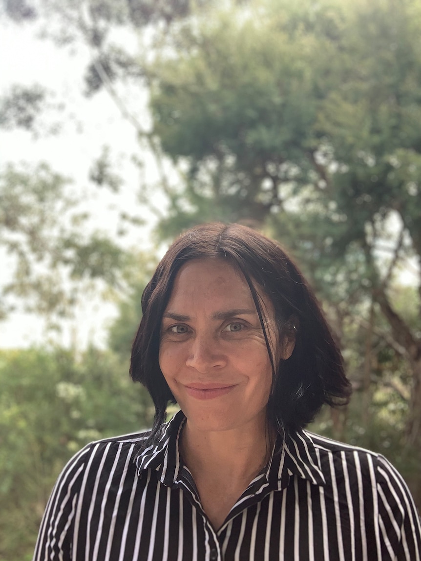 A woman with shoulder length dark wears monochrome vertical striped button up shirt, behind her are lush trees on overcast day.