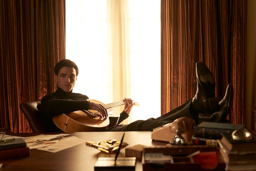 Jacob Elordi in character as Elvis Presley, holding a guitar, wearing a black jumper and his feet up on a desk