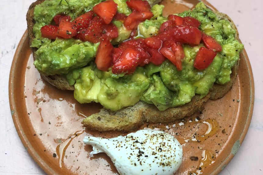 A brown plate is seen on top of a pink tabletop. There are two slices of sourdough on it, with smashed avocado and strawberries.