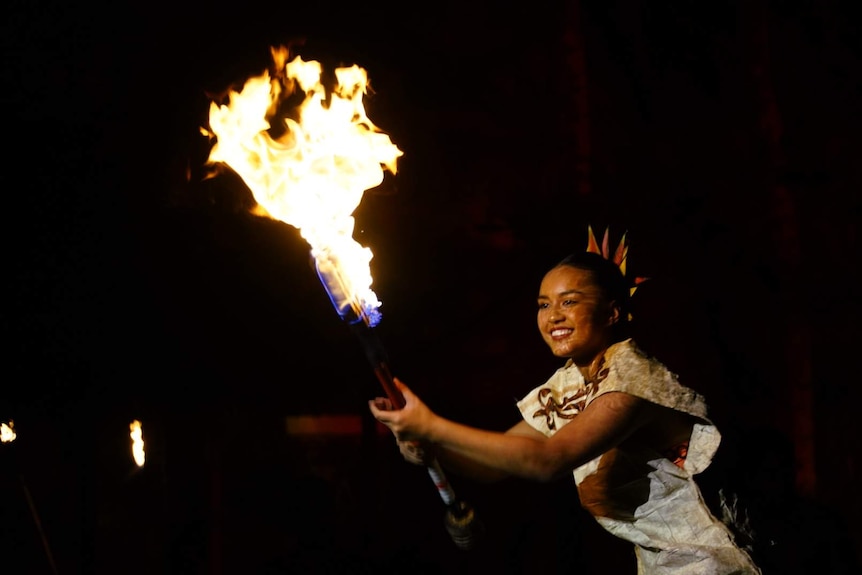 A woman in costume smiles swinging a long knife lit with large yellow flames.