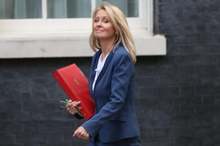 Esther McVey wears a blue suit and holds a red folder with a crown on it. She looks to camera as her blonde hair flows behind.