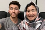 A man in a grey T-Shirt takes a selfie with his mother, who is wearing a hijab with grey and pink spots.