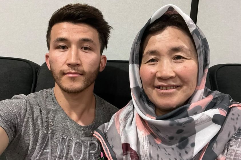 A man in a grey T-Shirt takes a selfie with his mother, who is wearing a hijab with grey and pink spots.