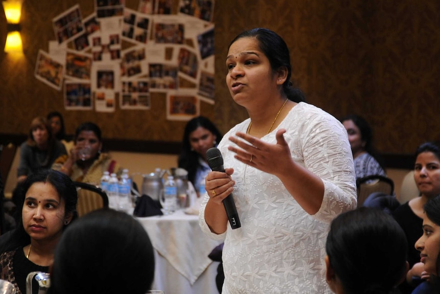 In a room full of people sitting around circular tables, a woman among them holding a microphone stands speaking.