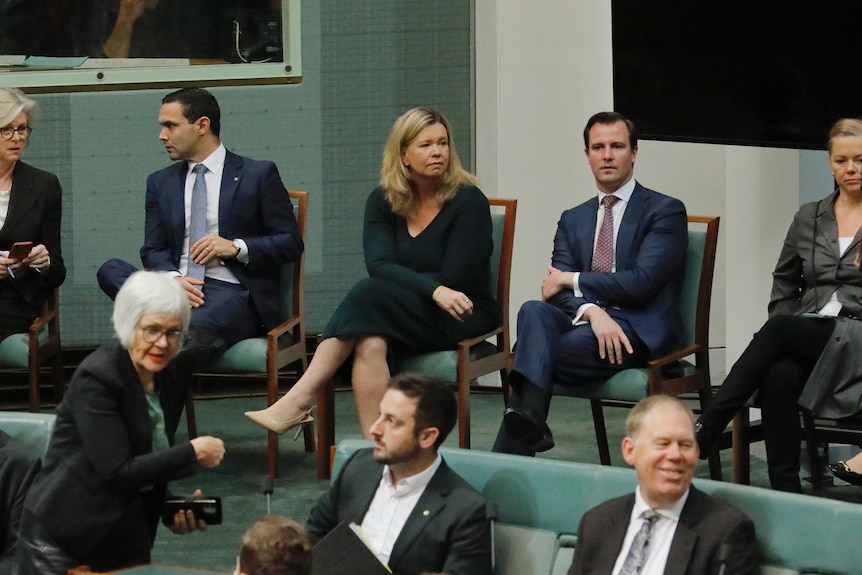 A blonde woman sitting in a chair surrounded by men in suits.