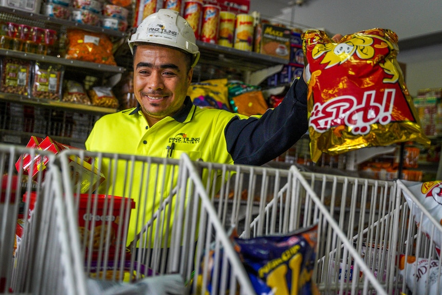 A man wearing a high vis shirt and hard hat reaches for a chip packet, smiling.