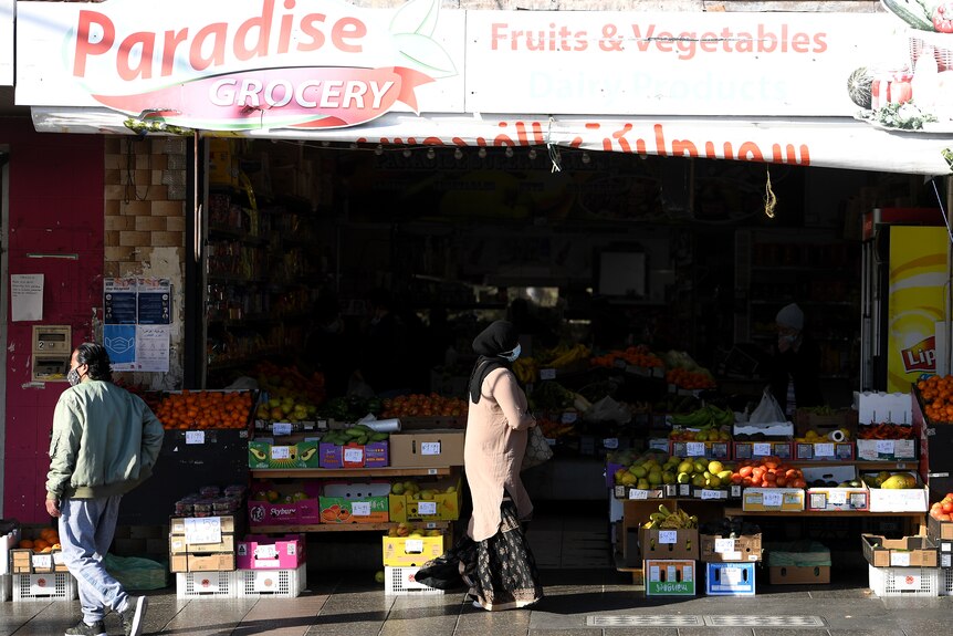A woman wearing a head covering and a mask stands in front of a fruit shop.