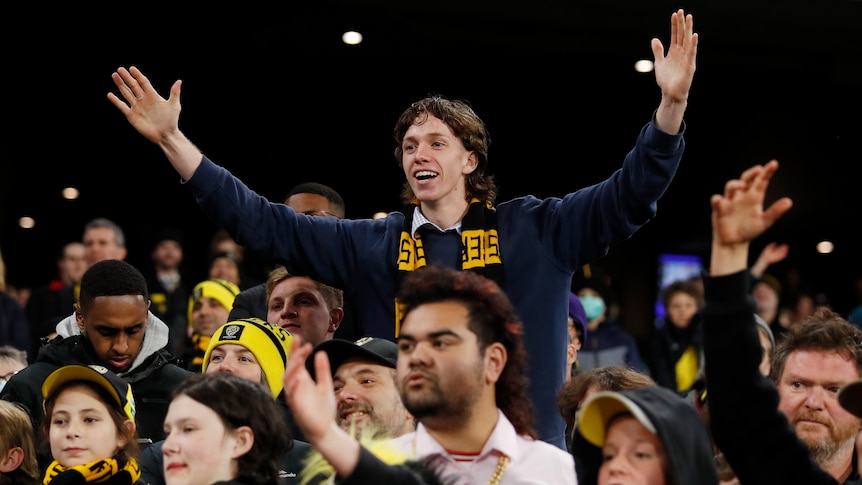 A young person with a yellow and black scarf holds his hands high in a crowd