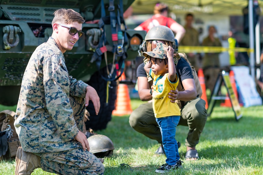 A boy wearing a yellow shirt and blue pants wears an army hat next to a marine
