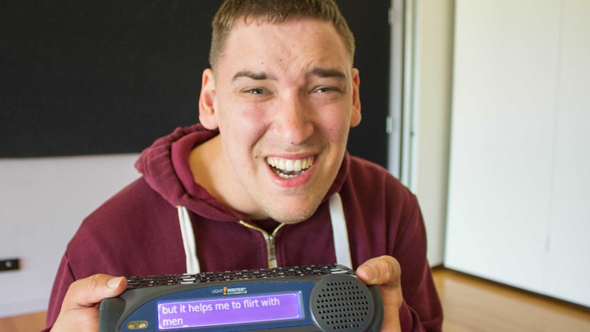 Thomas Banks smiling with his speech facilitator machine reading 'But it helps me to flirt with men'