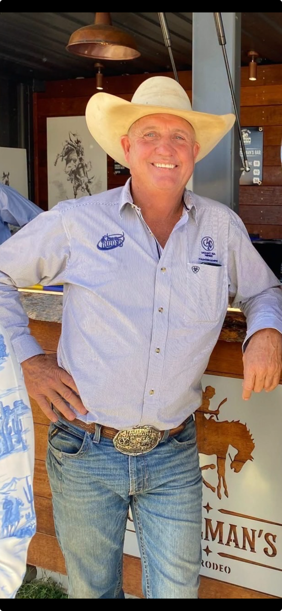 A man in a cowboy hat and rodeo shirt leans against a bar.