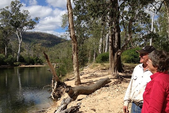 A grazier couple stand on the edge of their property overlooking a creek smiling.