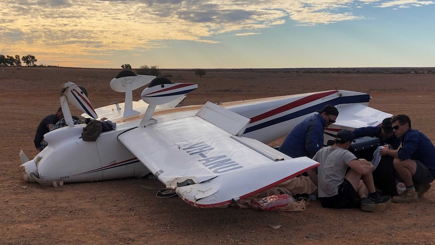 A light aircraft lying upside down on dirt while rescue workers attend to a figure lying under the wing.