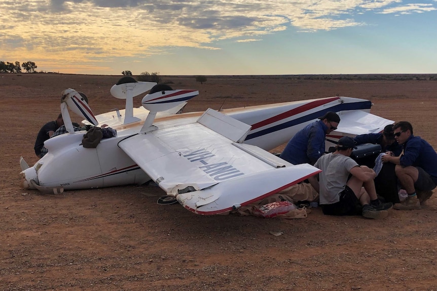 A light aircraft lying upside down on dirt while rescue workers attend to a figure lying under the wing.