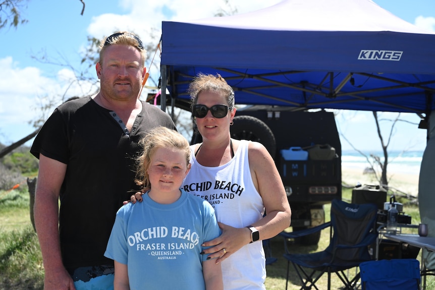 A man in a black shirt stands with his wife and daughter on a sunny day at their campsite.