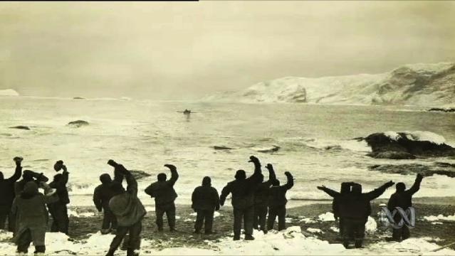 A group of men standing in Antarctica waving their hats in the air at a tiny boat in the distance