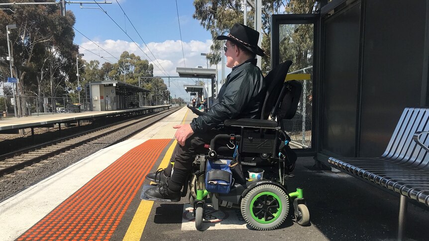 Brian Caccianiga waits behind the yellow train platform line in his wheelchair on a sunny day.