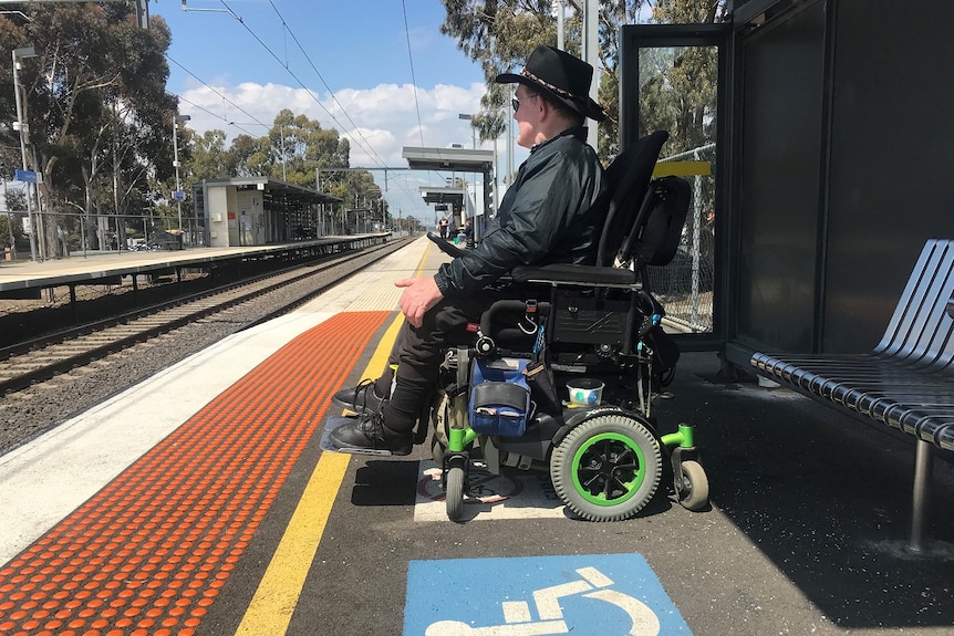 Brian Caccianiga waits behind the yellow train platform line in his wheelchair on a sunny day.