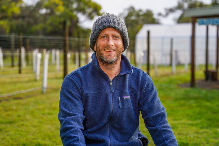 A man with a beard wearing a beanie and fleece jumper smiles for a photo outside.