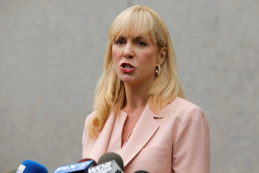 A blonde woman in a salmon-coloured suit speaks in front of microphones.