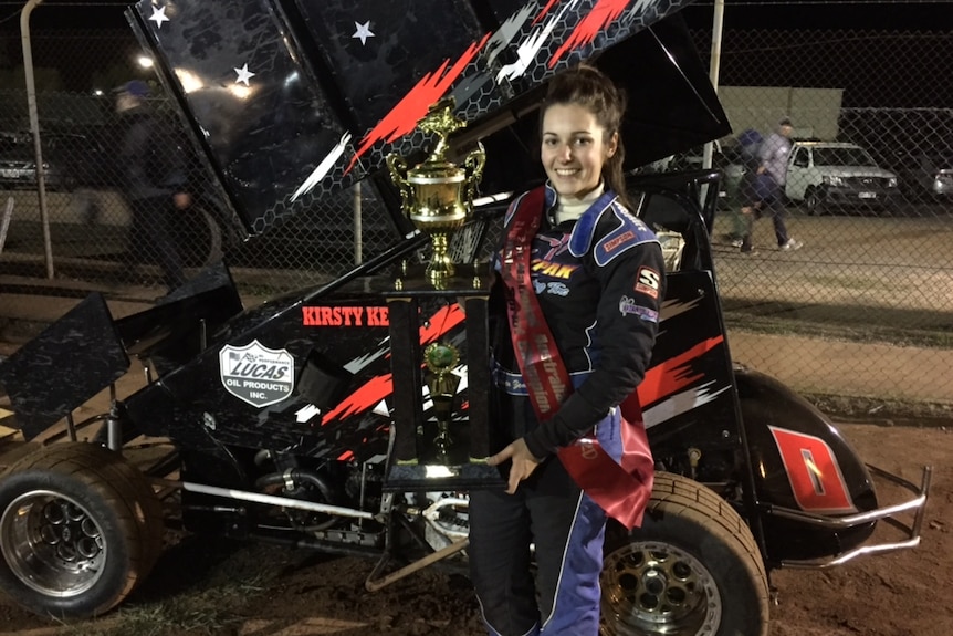 A young woman in a motorsport suit smiles holding a large gold trophy in front of a winged kart.