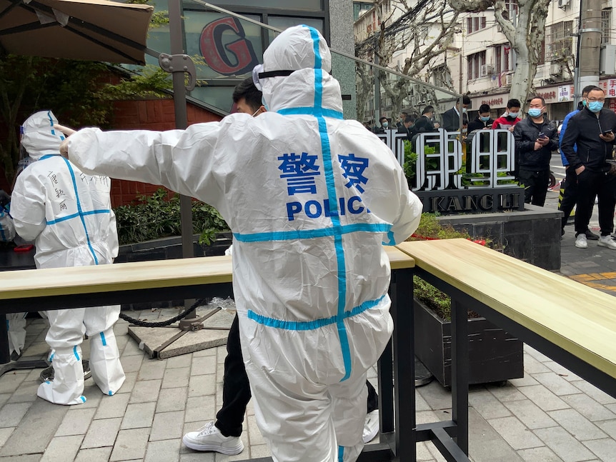 A man in all white protection gear with "Police" at the back, and people walk toward the direction he is giving to. 