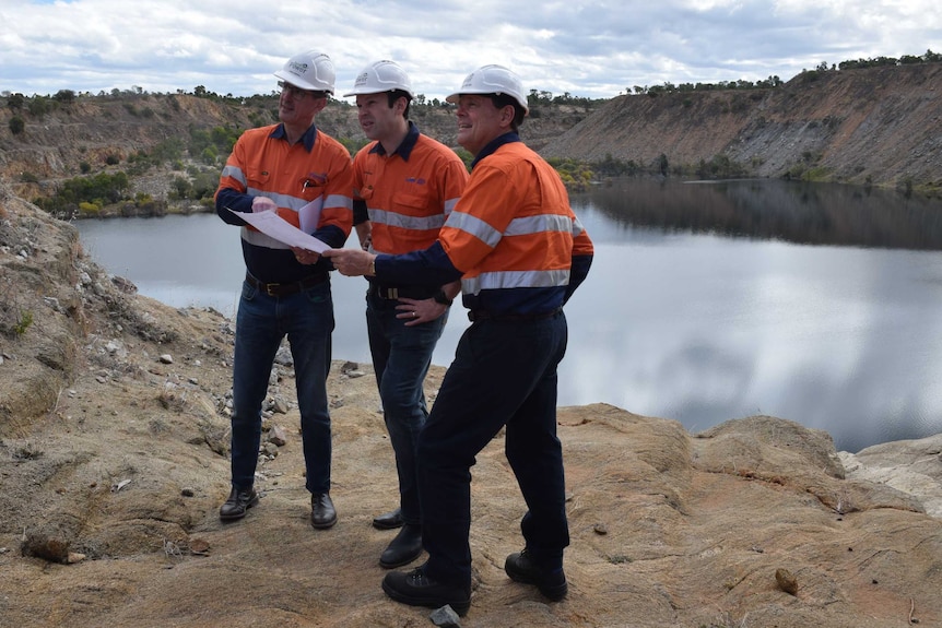 Three men wear hard hats and high vis holding a piece of paper with the mine pit behind them