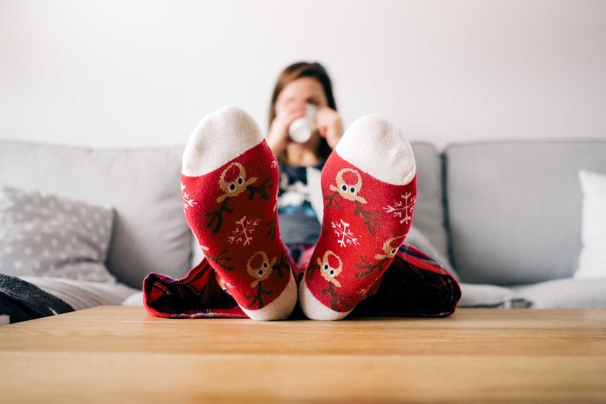 A woman sitting on a couch drinking from a mug with her feet in close up wearing festive socks