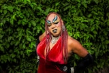 Colour photograph of drag act Nova Gina posing in front of a wall of green vines.