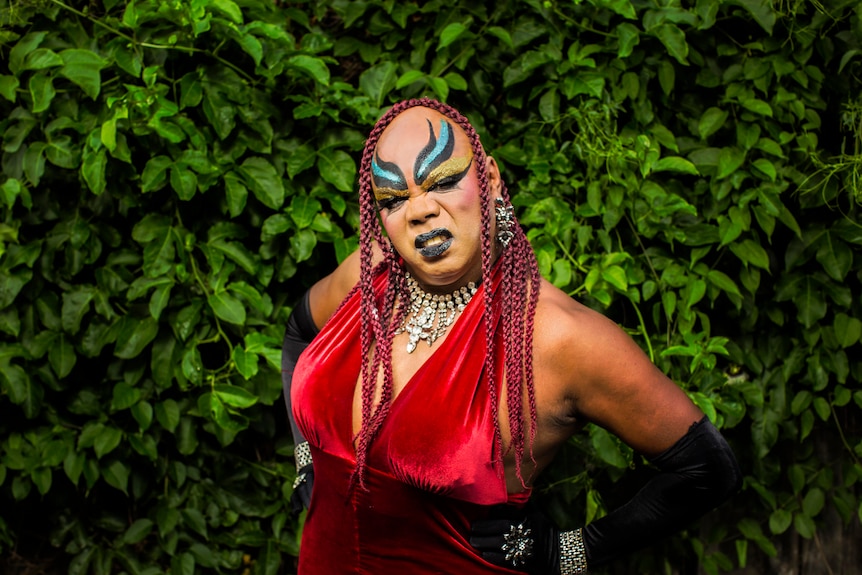 Colour photograph of drag act Nova Gina posing in front of a wall of green vines.