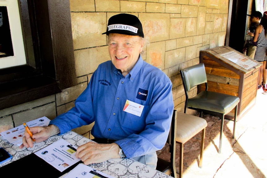 A man sits at a desk outside an old brick building, wearing a hat reading 'telegrapher'.