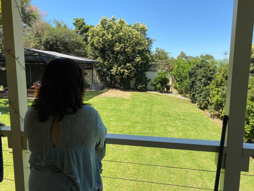A woman stands looking out over her backyard from a balcony on a sunny day.