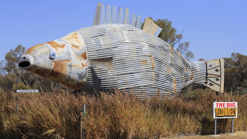 A giant metal fish made out of scrap and junk sits above bushes.