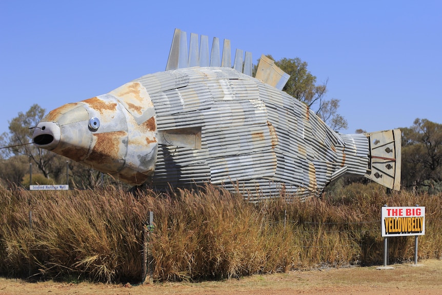 A giant metal fish made out of scrap and junk sits above bushes.