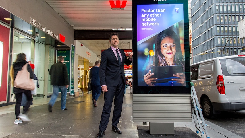 Councillor Nicholas Reece stands beside a large electronic billboard displaying a Telstra advertisement on a CBD footpath.