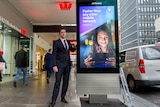 Councillor Nicholas Reece stands beside a large electronic billboard displaying a Telstra advertisement on a CBD footpath.