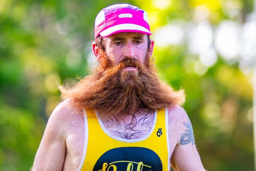 A man with a brown, ginger beard wearing a pink cap and yellow running singlet.