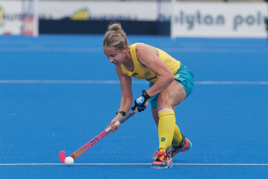 Female hockey player in green and gold Australia uniform bends over to hit a white ball with a wooden hockey stick.