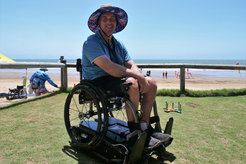 A white man smiling, sitting in a wheelchair, wearing a sunhat, in front of a sunny beach. 