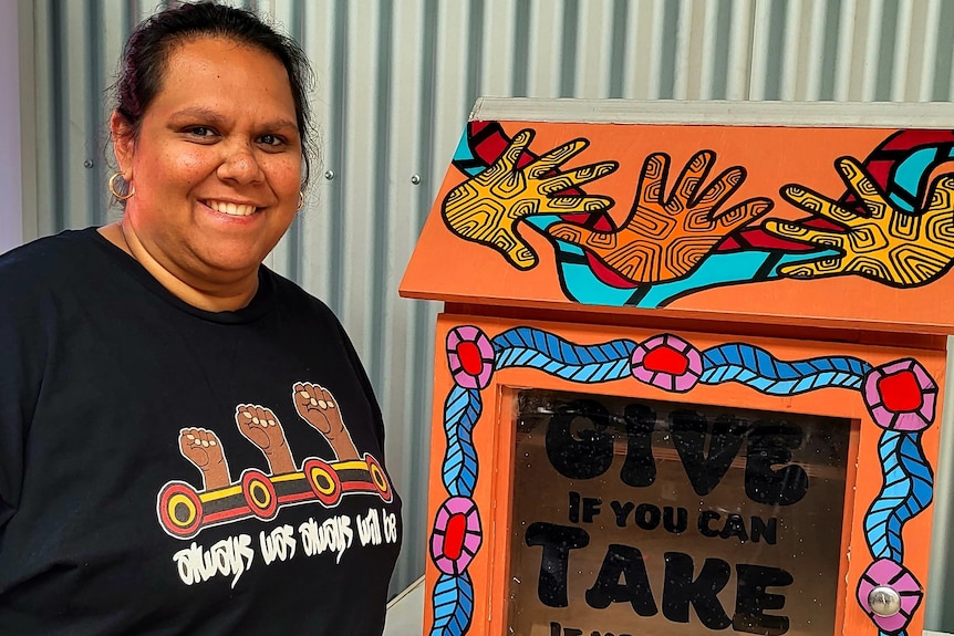 A female indigenous artist smiling stands next to a colourful wooden pantry box