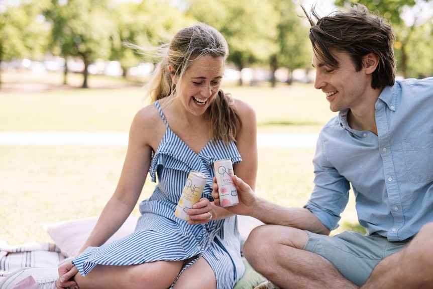 A woman and men sittting in a park drinking canned wine.