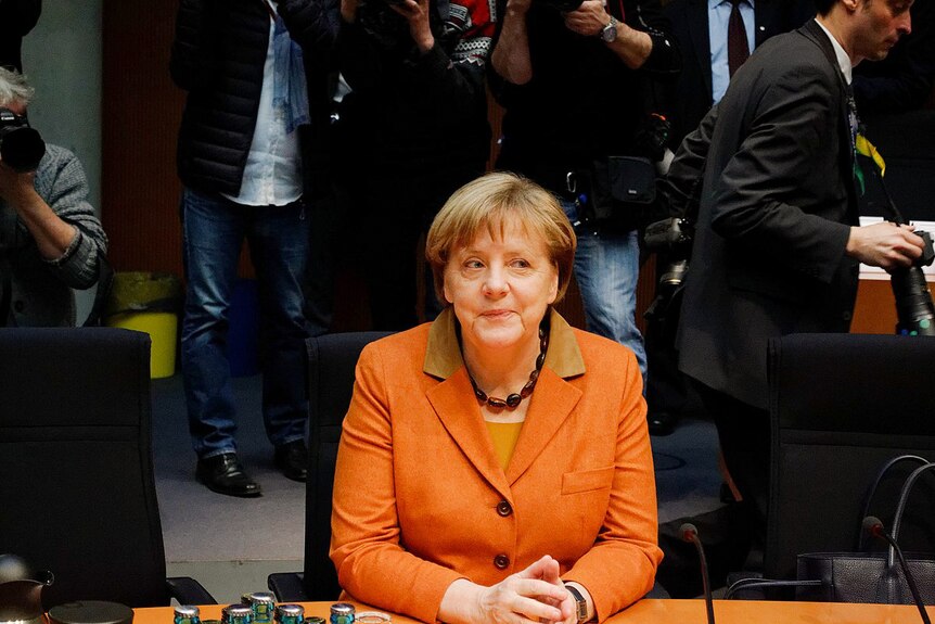 Angela Merkel in an orange blazer and wearing a black necklace smiles while sitting at a desk as people take photos of her.