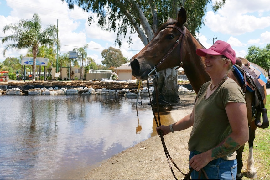 A woman stands next to her horse at a park in front of floodwaters lappiong at sandbags protecting a caravan park.