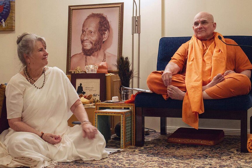 A woman in a white robe sits on the floor beside a man in orange clothing who sits on a low chair.