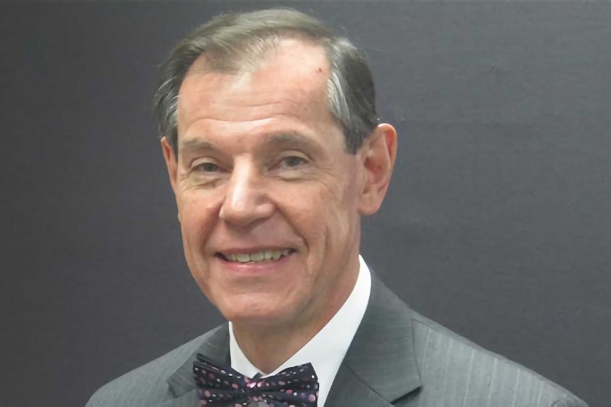 A man smiles at the camera in front of a grey wall