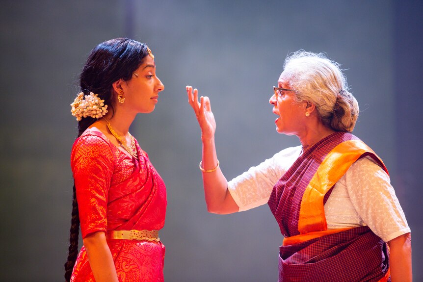 A young Sri Lankan woman in a red sari and an older Sri Lankan woman in a white and maroon sari, holding her hand up