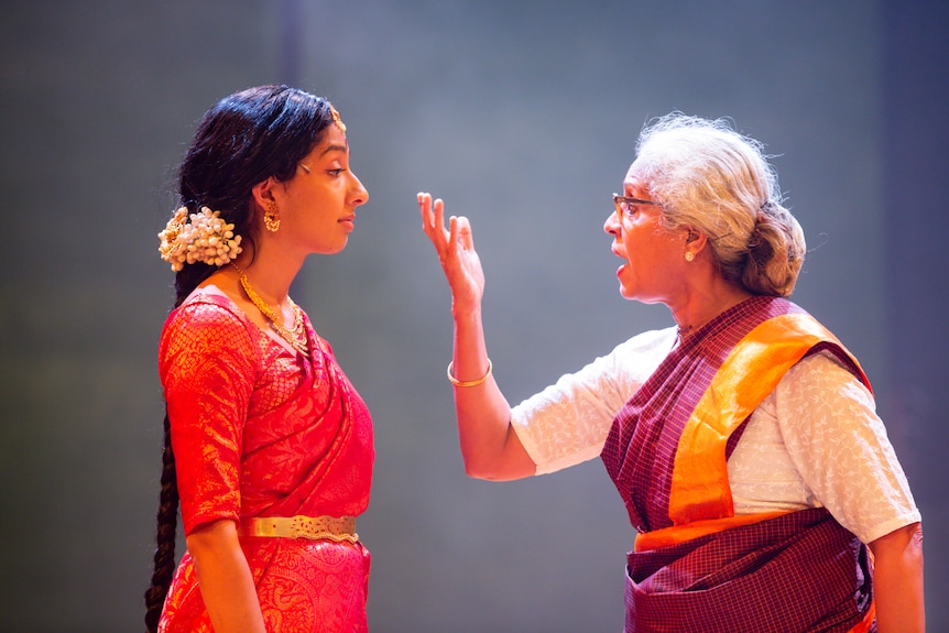 A young Sri Lankan woman in a red sari and an older Sri Lankan woman in a white and maroon sari, holding her hand up