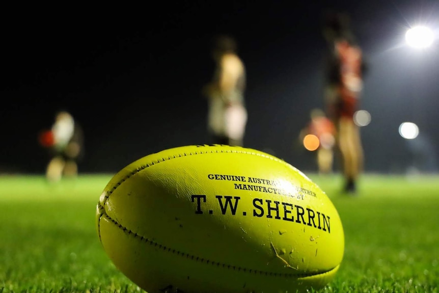 A small florescent green football lies on a football oval under lights in the foreground as men train in the background.